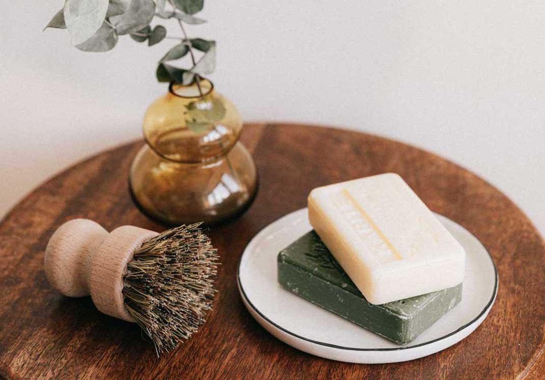 Two bars of soap resting on a small plate, plus a brush and small vase with eucalyptus sitting on a round stool in a short term rental bathroom.