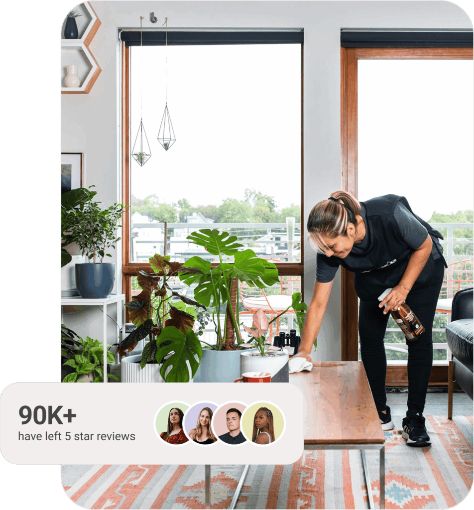 A professional cleaner wipes down a wooden coffee table in a bright, modern living room filled with green houseplants and natural light from large windows. A text overlay in the corner reads &ldquo;90K+ have left 5-star reviews,&rdquo; accompanied by profile icons of four diverse people representing satisfied Spruce customers.